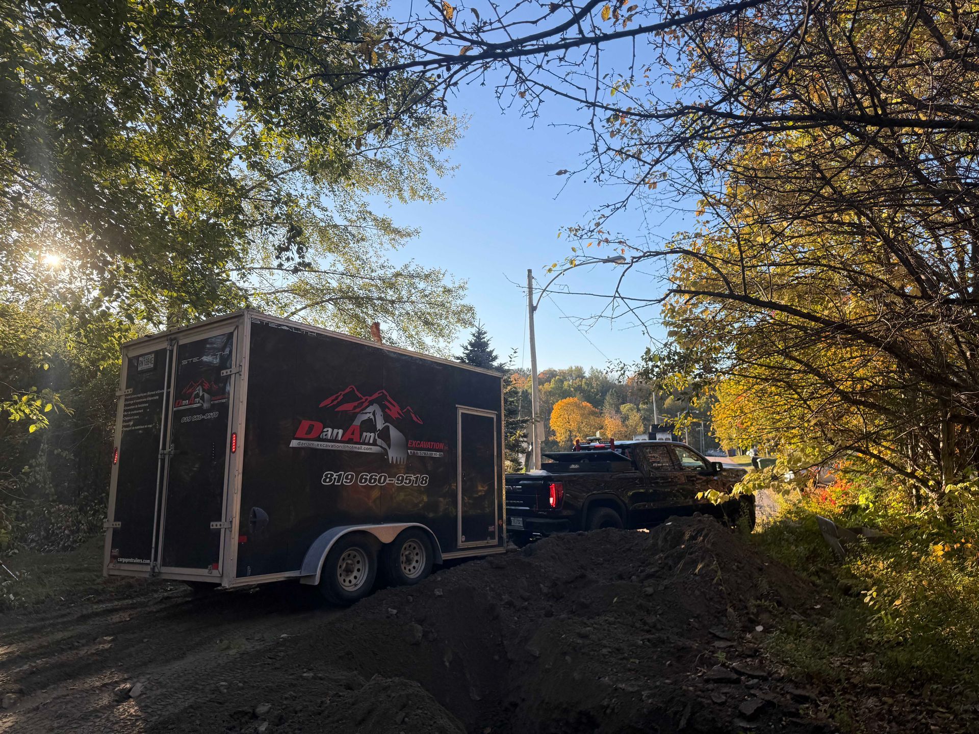 Une remorque noire arborant un logo est attelée à une camionnette sombre, garée sur un chemin de terre à côté d'un tas de terre.