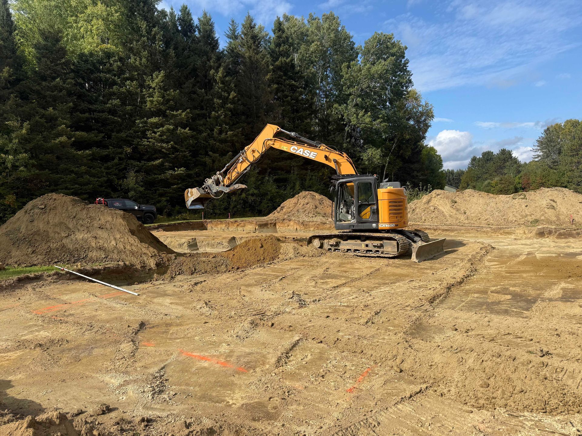 Une pelleteuse jaune trône au milieu d'un vaste chantier sablonneux, avec des tas de terre et une forêt en arrière-plan.
