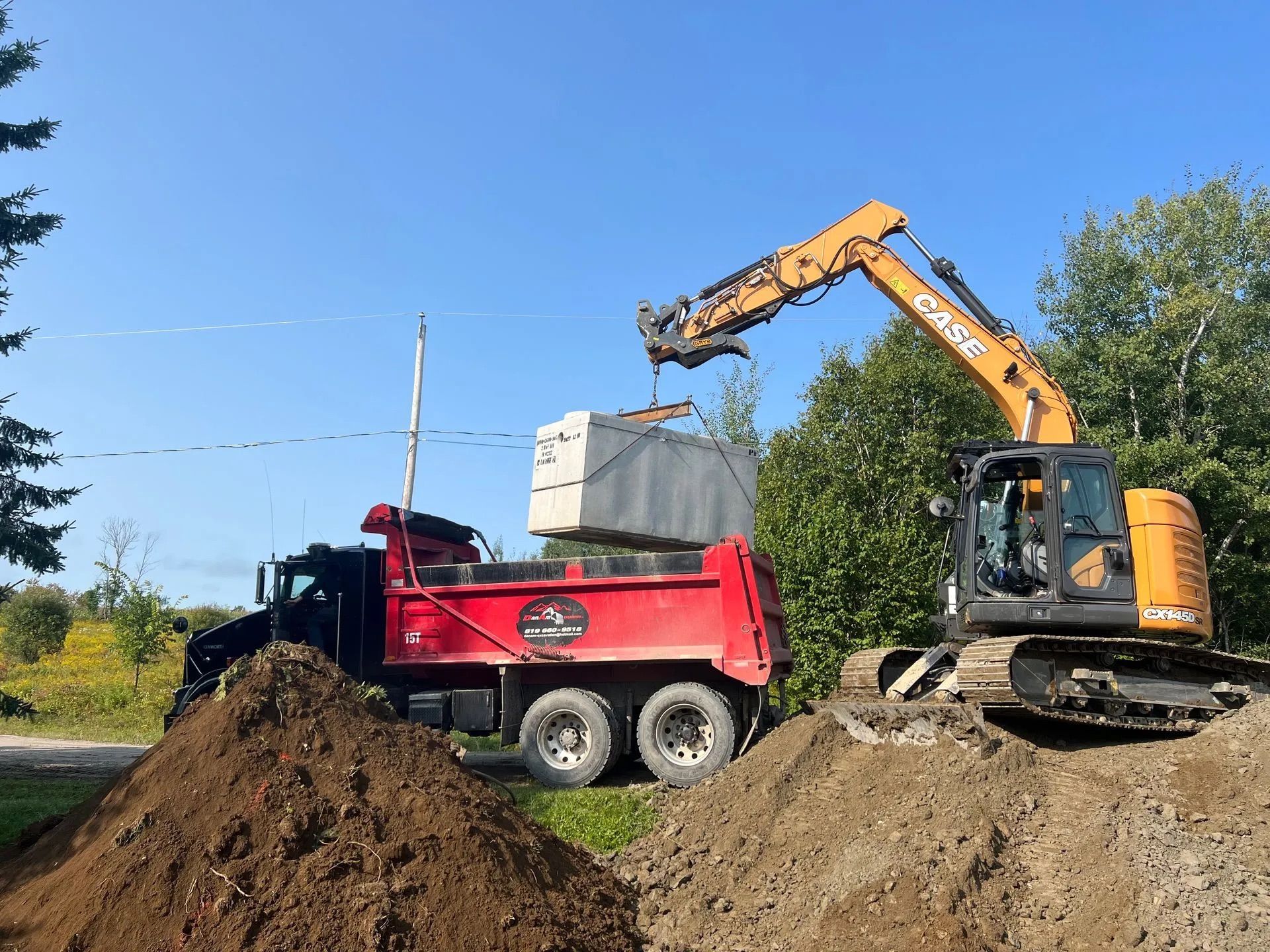 Une pelleteuse orange soulève un bloc de béton et le dépose dans un camion-benne rouge stationné entre deux gros tas de terre, sous un ciel bleu.
