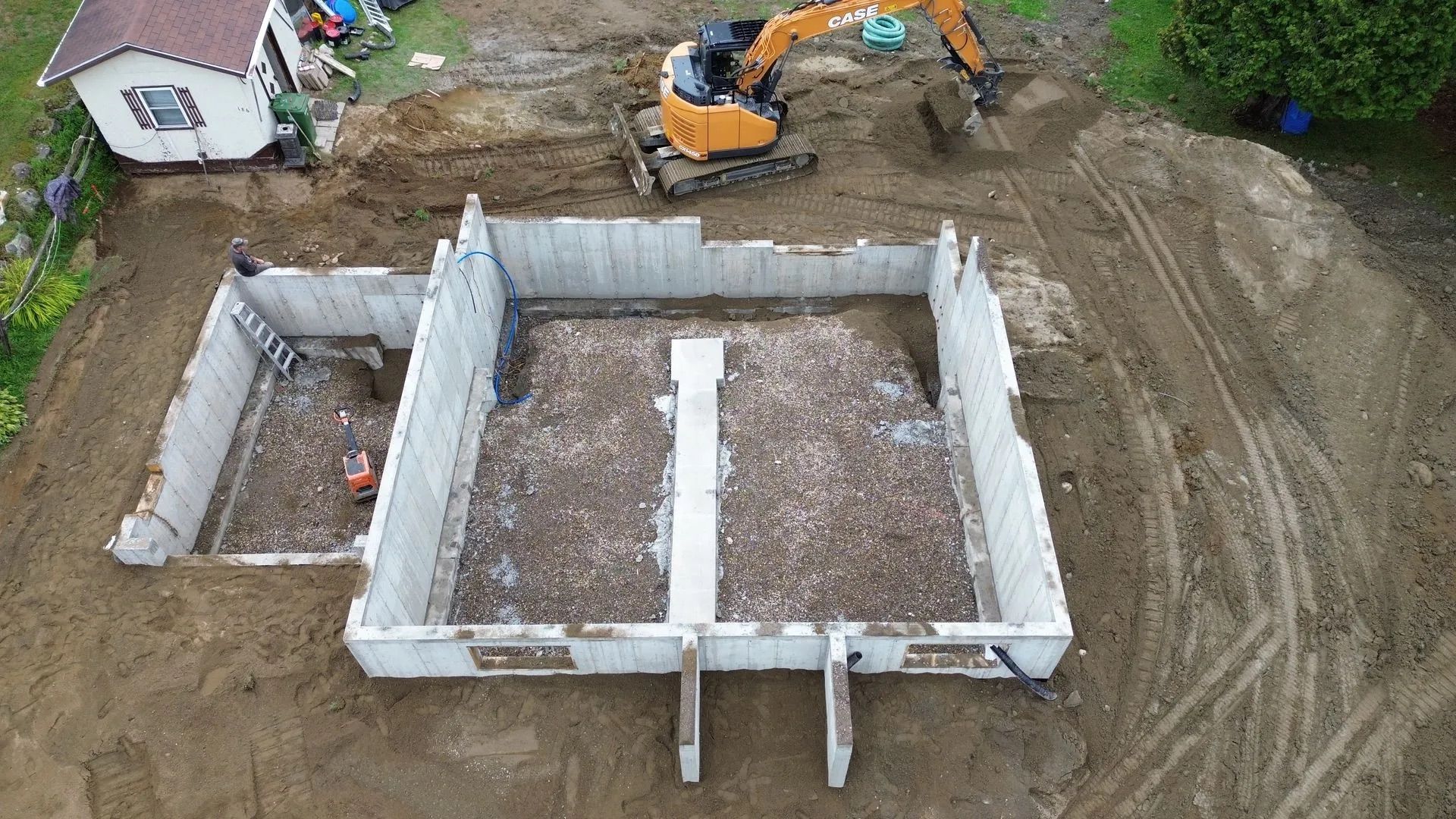 Vue aérienne des fondations d'une maison en béton en construction, avec une excavatrice à proximité, sur un terrain vague en terre battue.