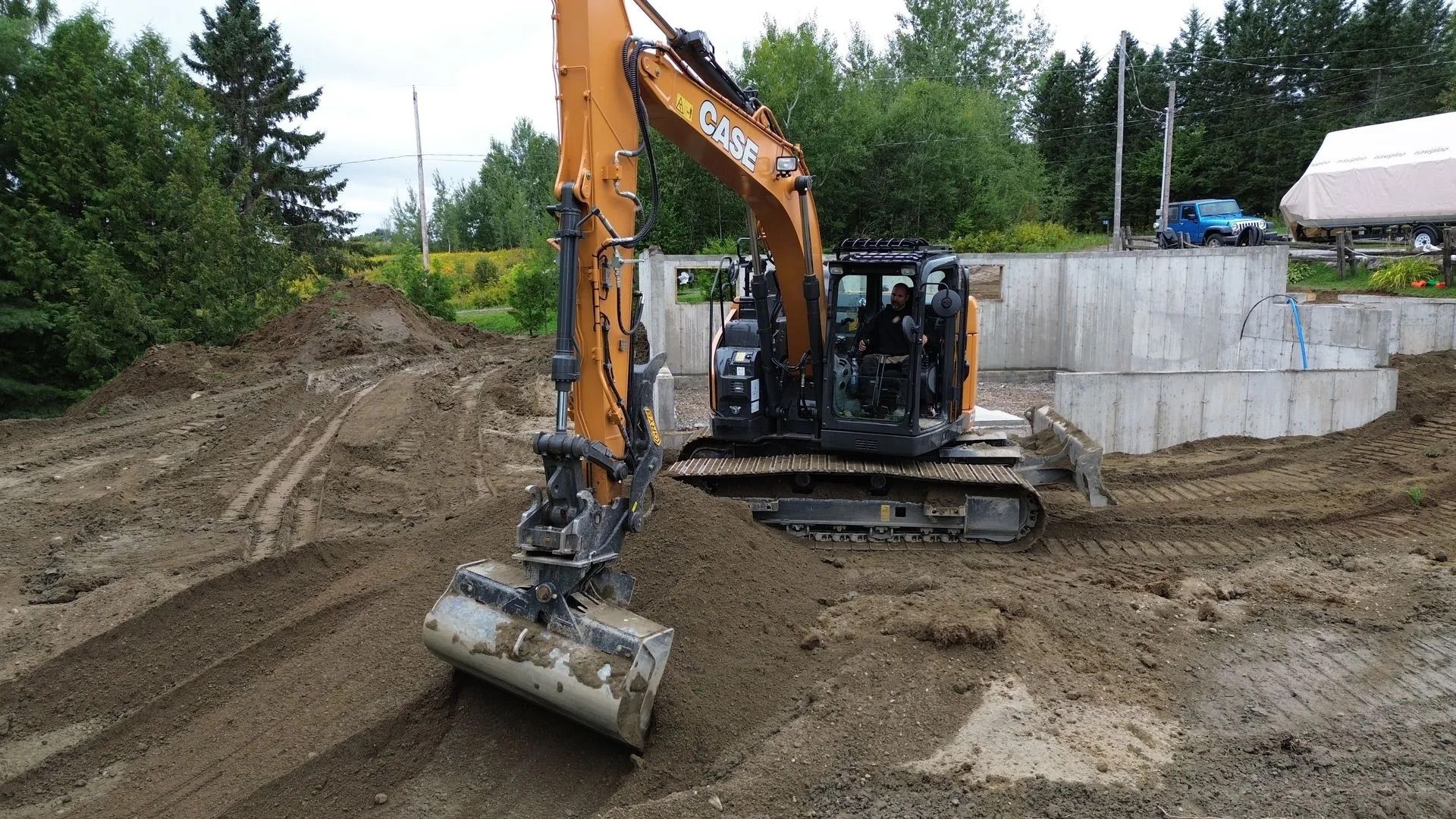 Une pelle mécanique Case orange, godet abaissé, travaille sur un chantier de construction en terre battue, à côté de fondations en béton.