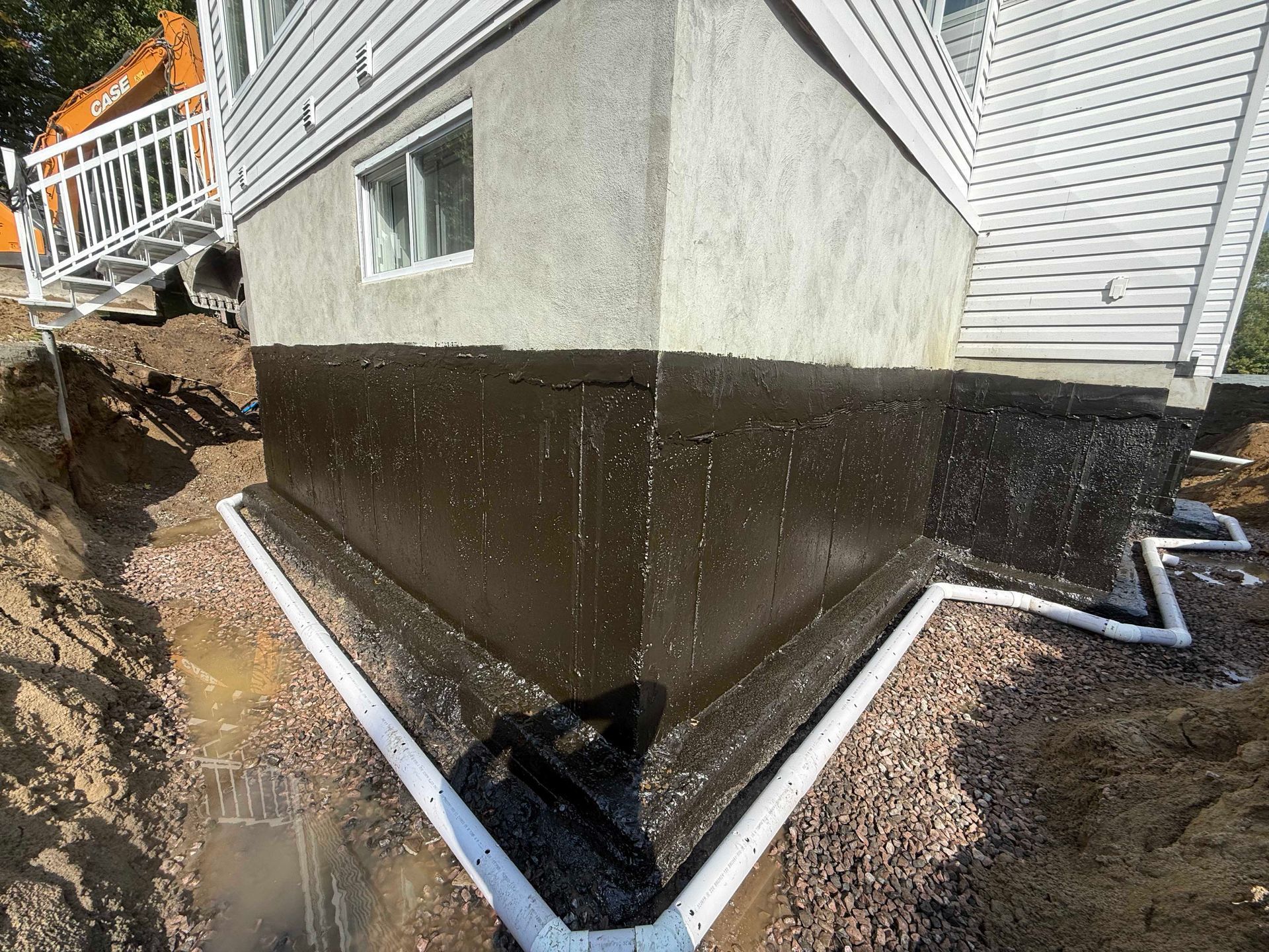 Les fondations d'une maison sont recouvertes d'un revêtement imperméable noir neuf et un tuyau de drainage blanc est installé autour de la base.