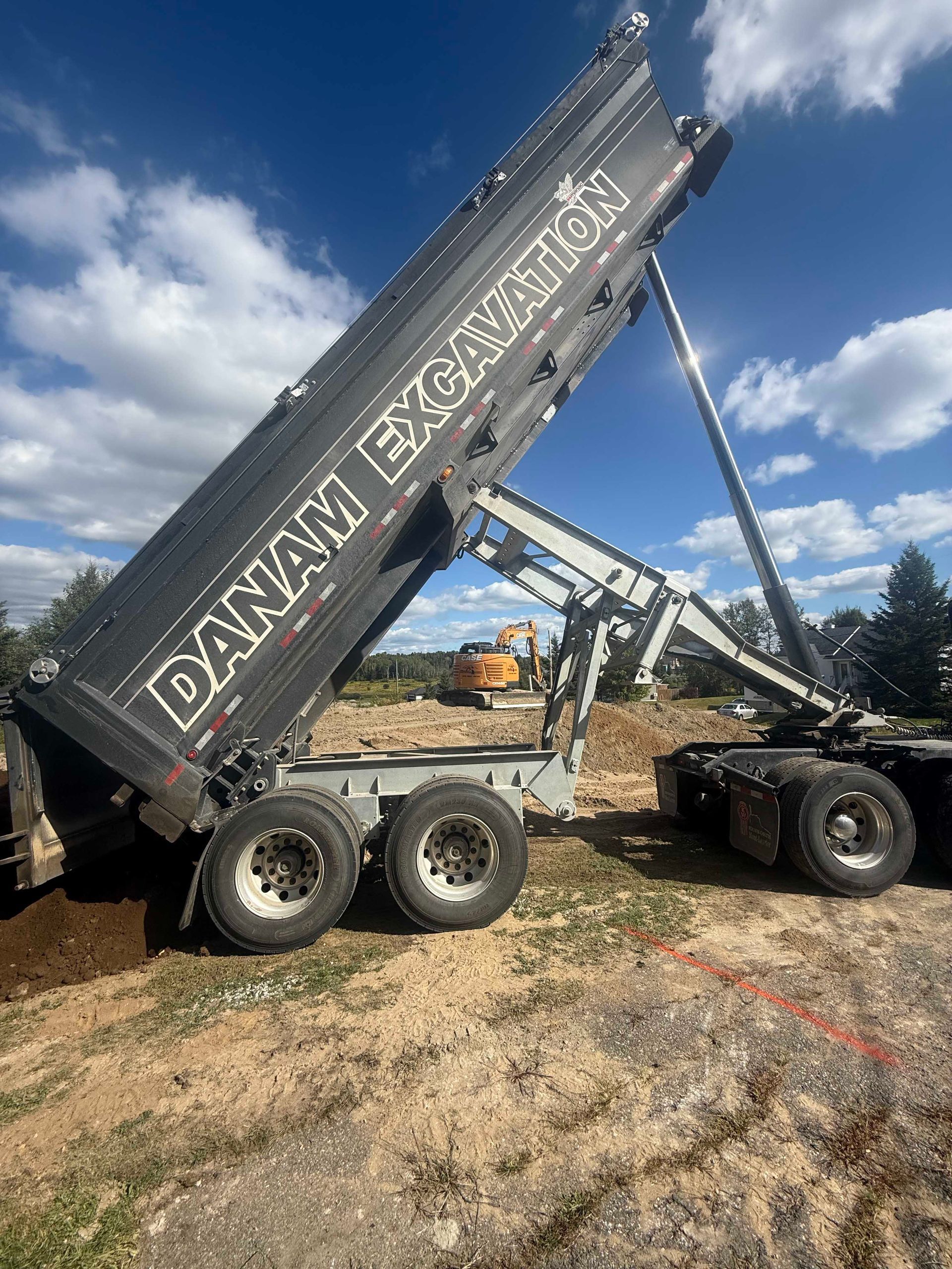 Un camion-benne noir de la société Danam Excavation est stationné sur un terrain vague, sa benne entièrement levée pour déverser de la terre.