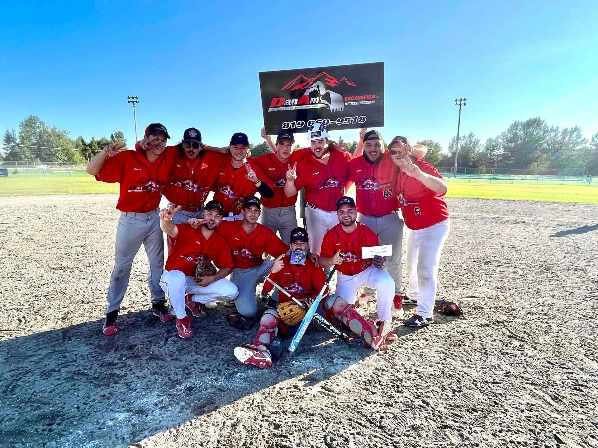 Une équipe de softball en maillots rouges et pantalons blancs célèbre sa victoire en brandissant une pancarte sur un terrain en terre battue.