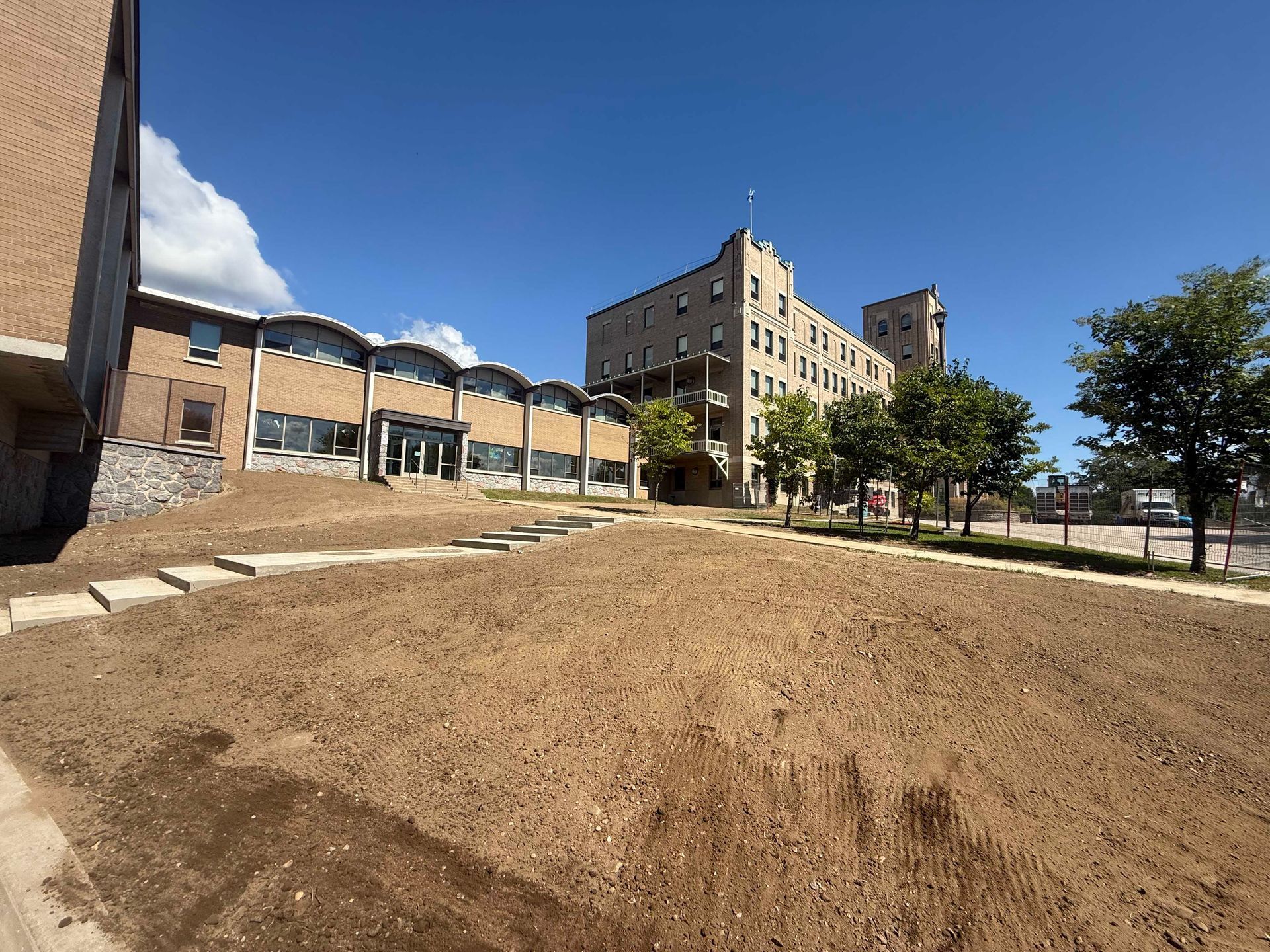 Sous un ciel bleu clair, un bâtiment moderne au toit incurvé se dresse à côté d'un grand immeuble en briques beiges, au milieu d'une pelouse en terre battue.
