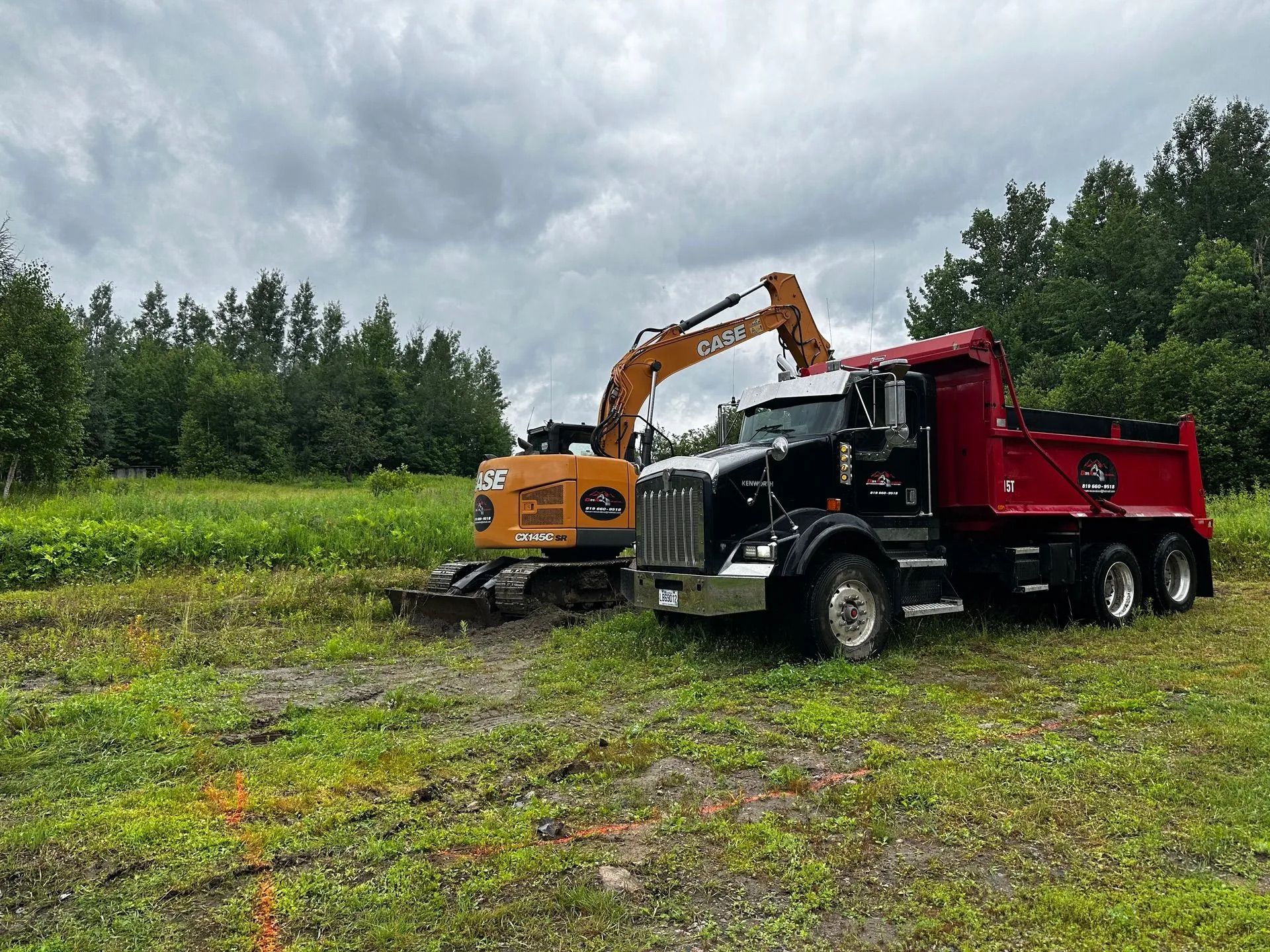 Une pelleteuse orange charge de la terre dans un camion-benne rouge sur un champ herbeux sous un ciel nuageux.