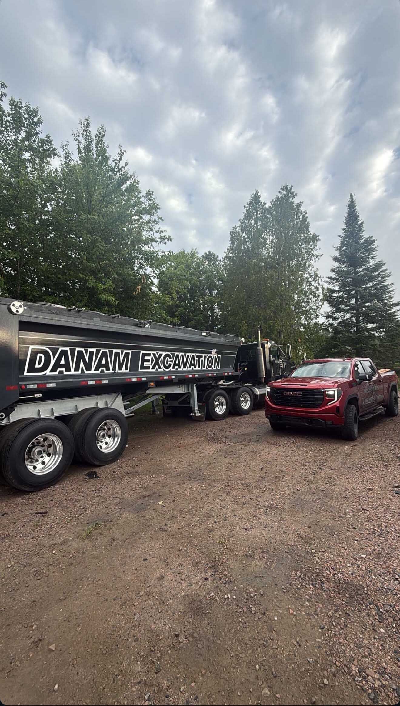 Un pick-up rouge est garé à côté d'une grande benne noire portant l'inscription « DANAM » sur un terrain gravillonné près d'arbres.