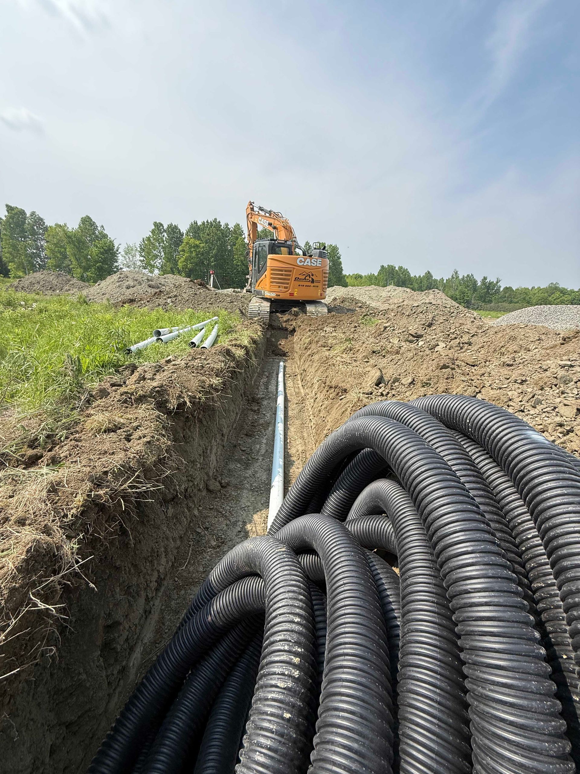 Une excavatrice garée dans une profonde tranchée par une journée ensoleillée, avec une grande bobine de tuyau ondulé noir au premier plan.