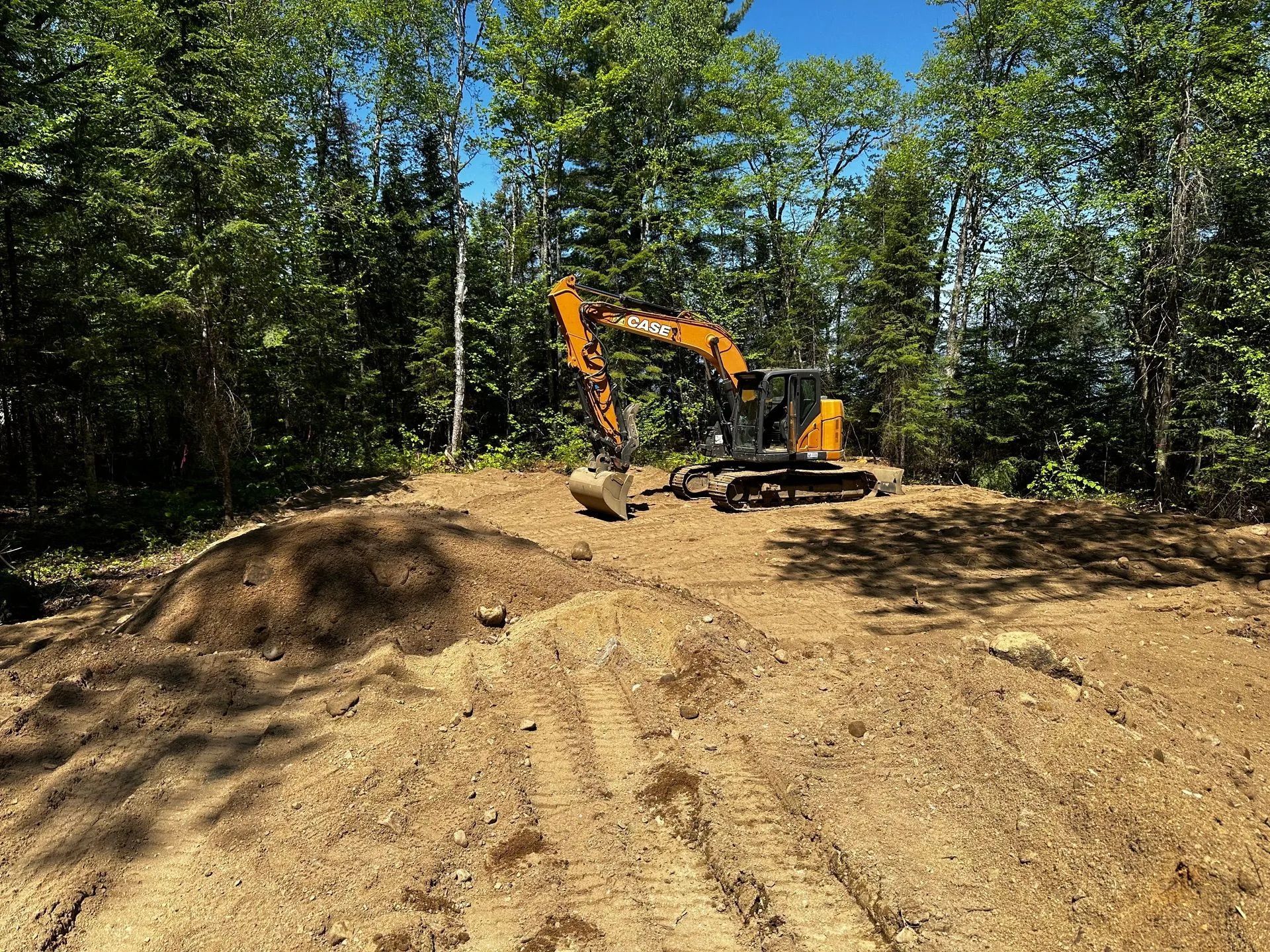 Une pelleteuse orange est posée sur un terrain vague recouvert de terre, entouré d'une forêt verdoyante et dense, sous un ciel d'un bleu limpide.