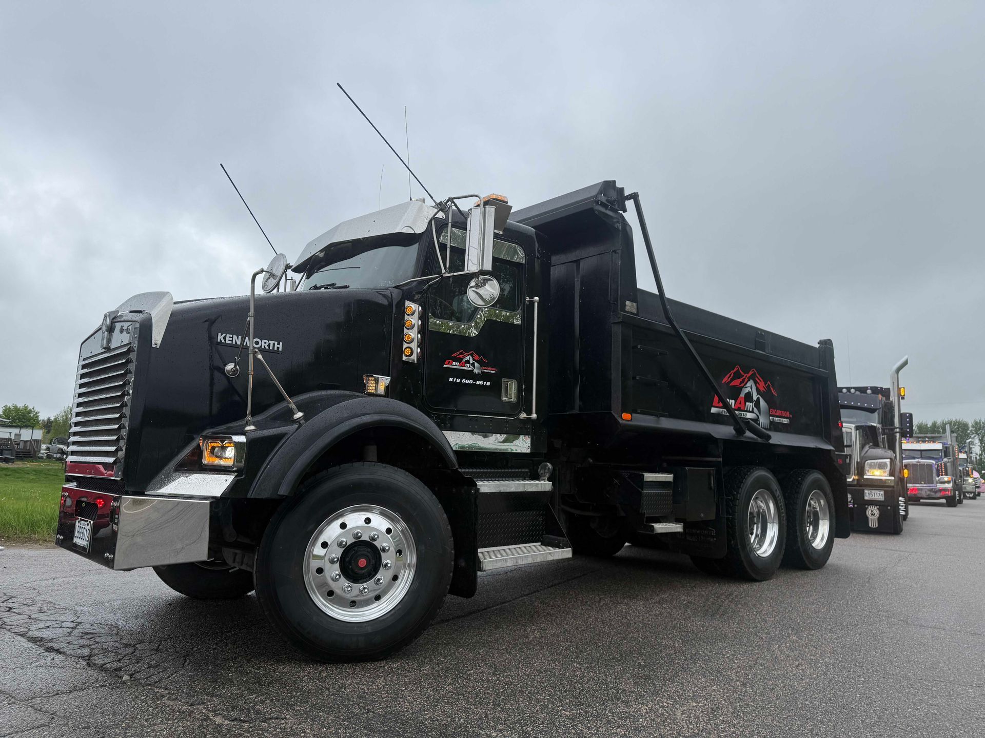Un élégant camion-benne Mack noir est stationné sur une surface asphaltée mouillée sous un ciel nuageux, avec d'autres camions visibles derrière lui.