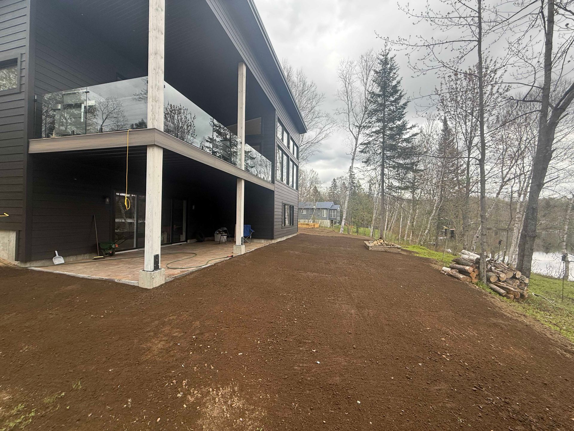 Une maison moderne aux panneaux sombres avec un balcon à balustrade en verre donnant sur une grande cour en terre fraîchement préparée.