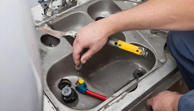 Person using a screwdriver to repair a sink drain in a stainless steel kitchen sink