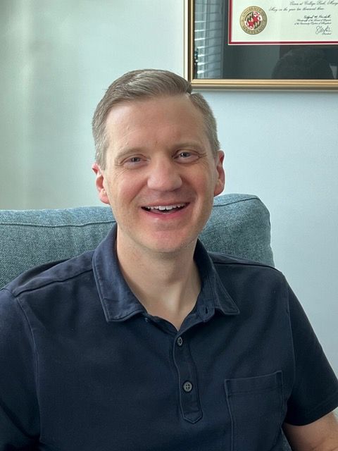A man in a blue shirt is sitting in a chair and smiling.
