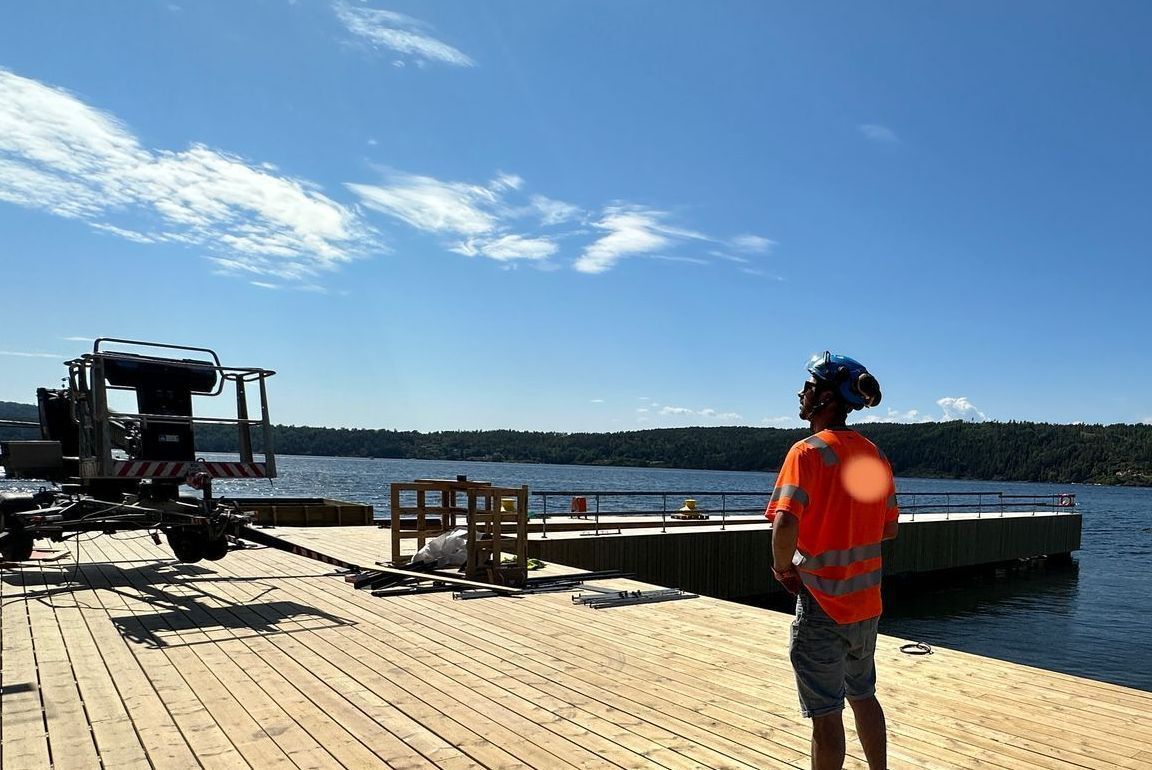 Man in orange vest on wooden dock, crane in foreground, blue sky and water.