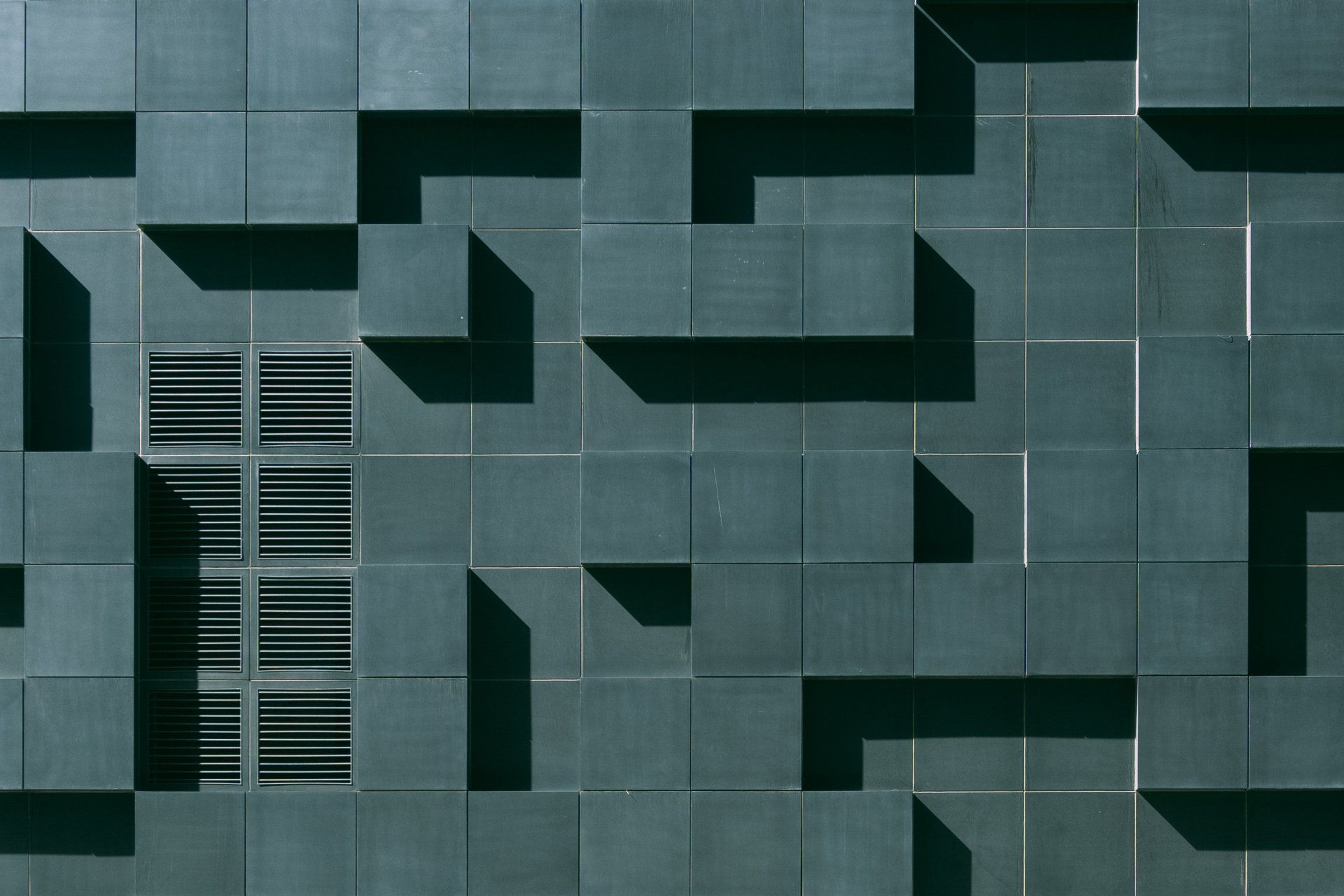 Facade with textured dark green square tiles, cast shadows. Ventilation vent.