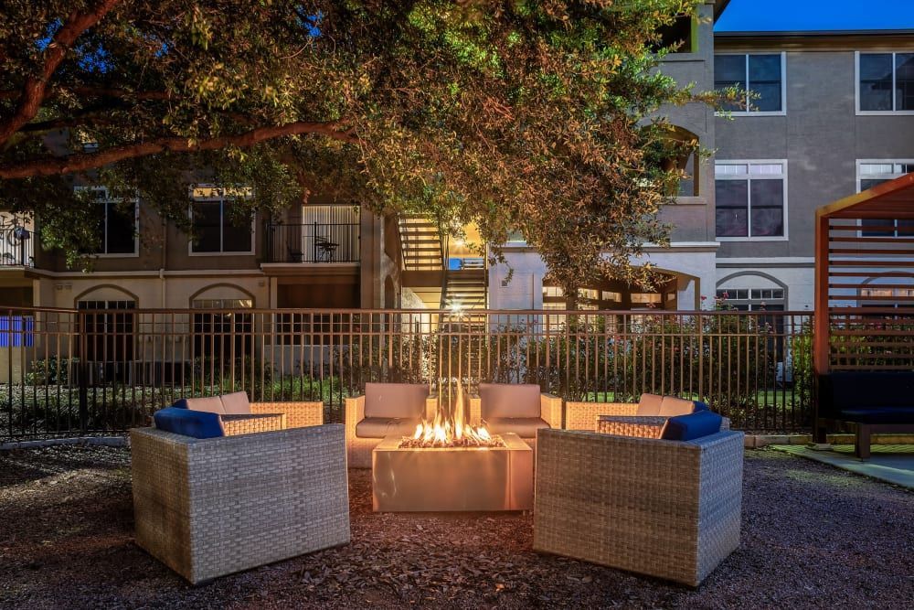 Evening view of a cozy outdoor seating area with a lit fire pit, surrounded by wicker armchairs with blue cushions, under the expansive branches of a large tree in front of a multi-story residential building at Marquis at Barton Trails in Austin, TX.