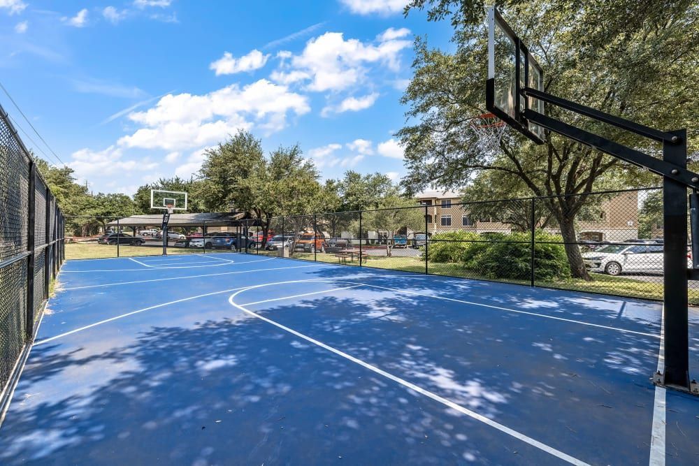 An outdoor basketball court with a vibrant blue playing surface and white markings. The court is surrounded by a chain-link fence, with basketball goals at either end. Trees and a parking lot with cars are visible in the background, under a clear blue sky at Marquis at Barton Trails in Austin, TX.