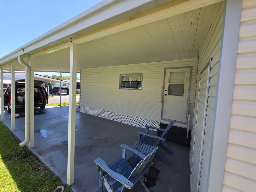 Covered porch with white posts, two blue chairs, and a door beside a mobile home.