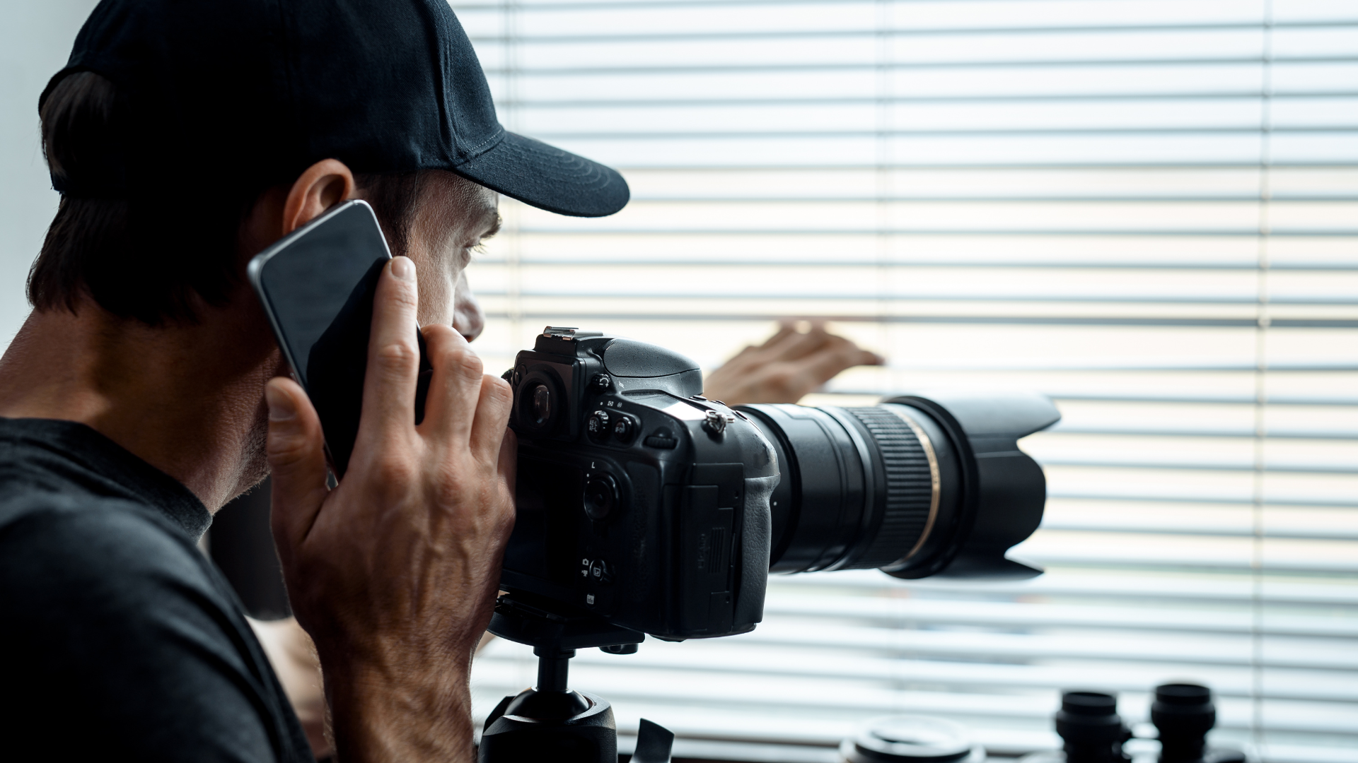 Person on phone looks through window blinds with a camera on a tripod.