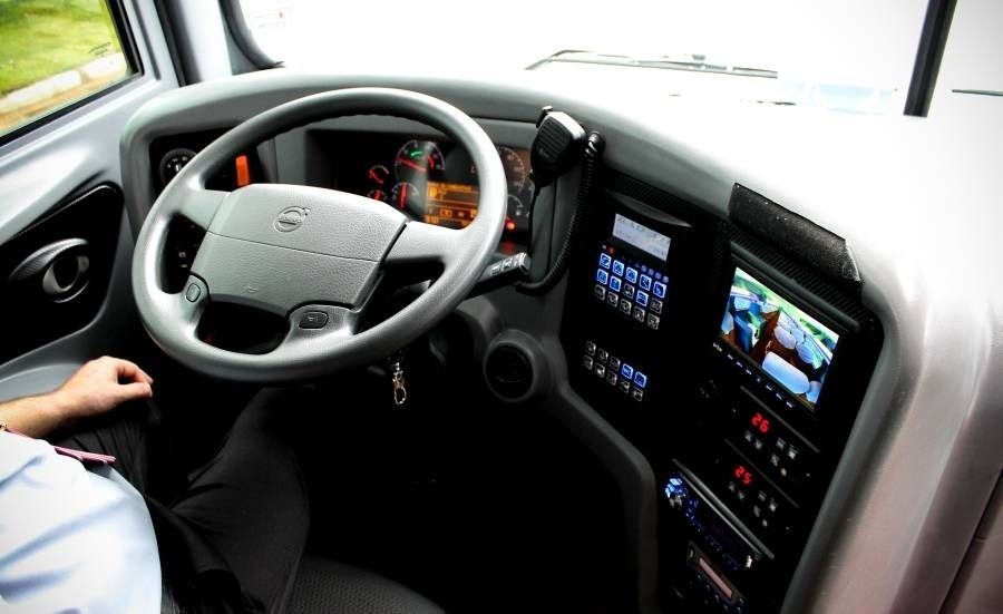 Interior do ônibus com motorista, volante e telas de exibição. Painel cinza e preto.
