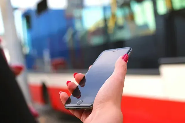 Mão segurando um smartphone com unhas vermelhas e ônibus desfocado ao fundo.