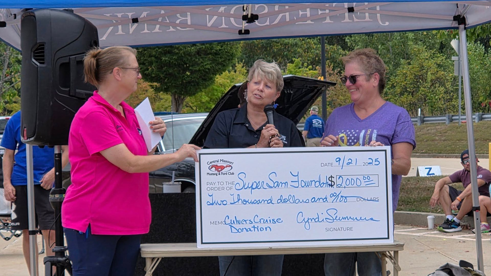Three people stand under a tent holding a large ceremonial check during an outdoor community event.
