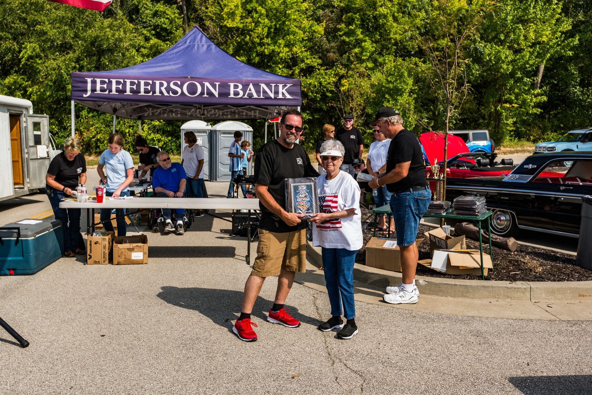 A person presents a trophy to another individual outdoors at a Jefferson Bank community event with cars parked nearby.