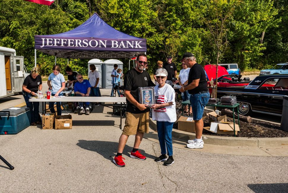 A person presents a trophy to another individual outdoors at a Jefferson Bank community event with cars parked nearby.