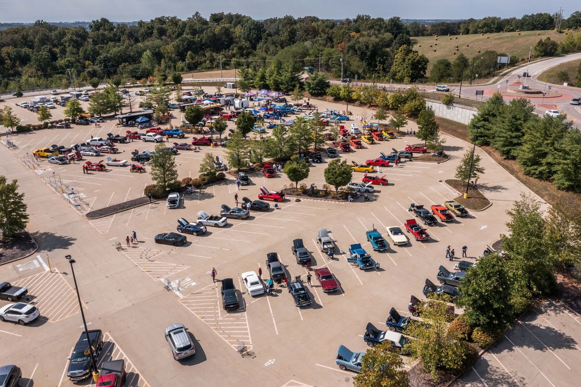 An aerial view of a car show with many vehicles parked in rows within an outdoor parking lot surrounded by trees.