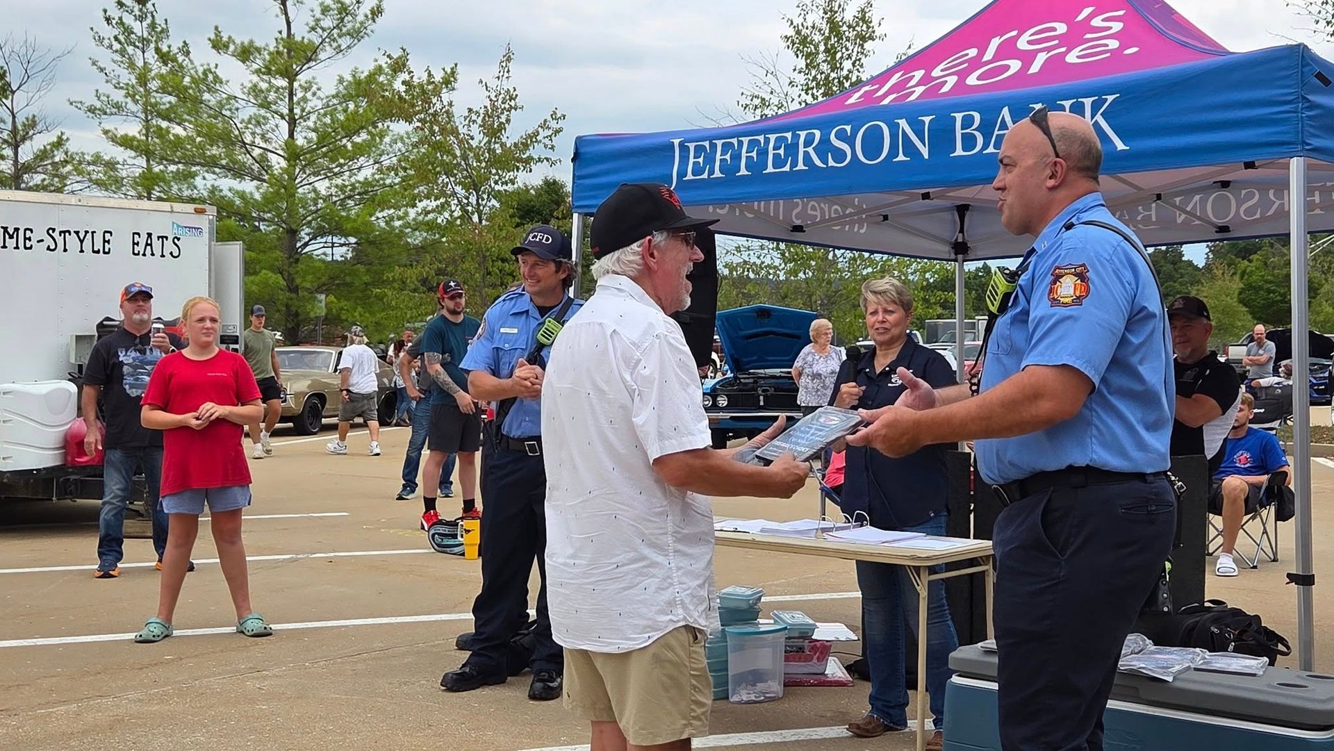 Two uniformed officials present an award to a person under a Jefferson Bank tent during an outdoor community event.
