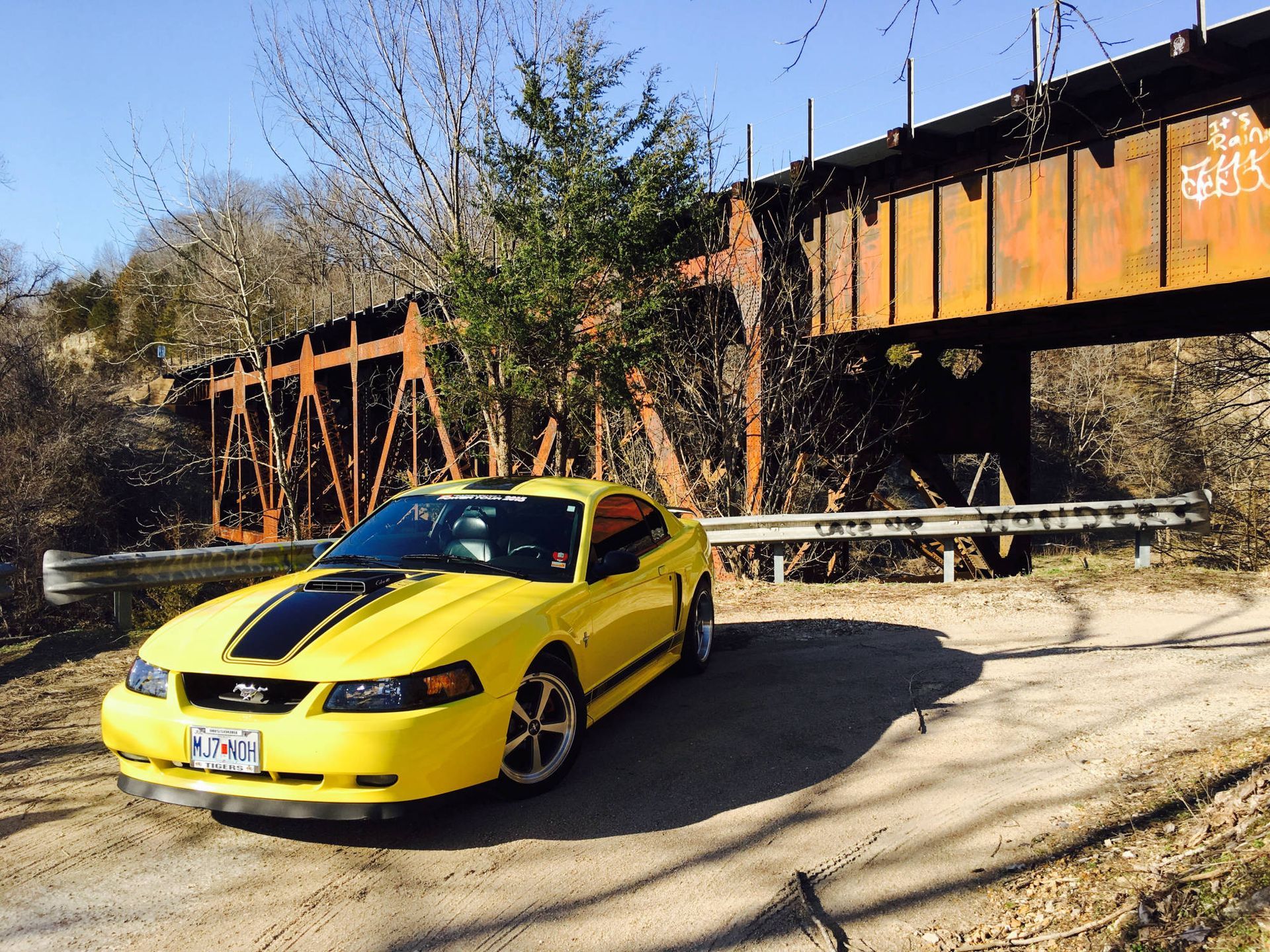 A bright yellow Ford Mustang with black racing stripes parked on a dirt road beneath a weathered, rusty metal bridge.
