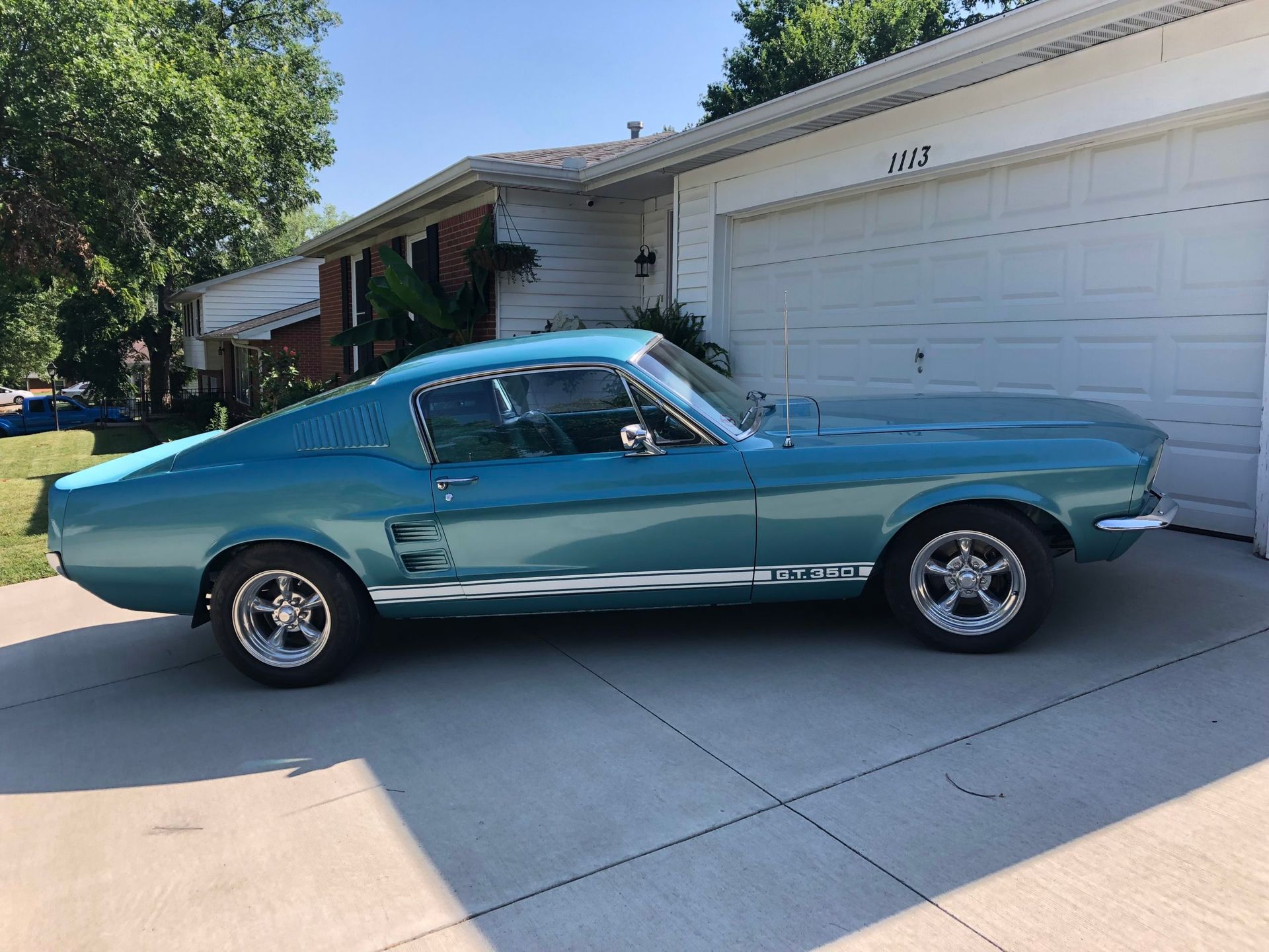 A teal classic Ford Mustang fastback parked in a suburban driveway in front of a white house.