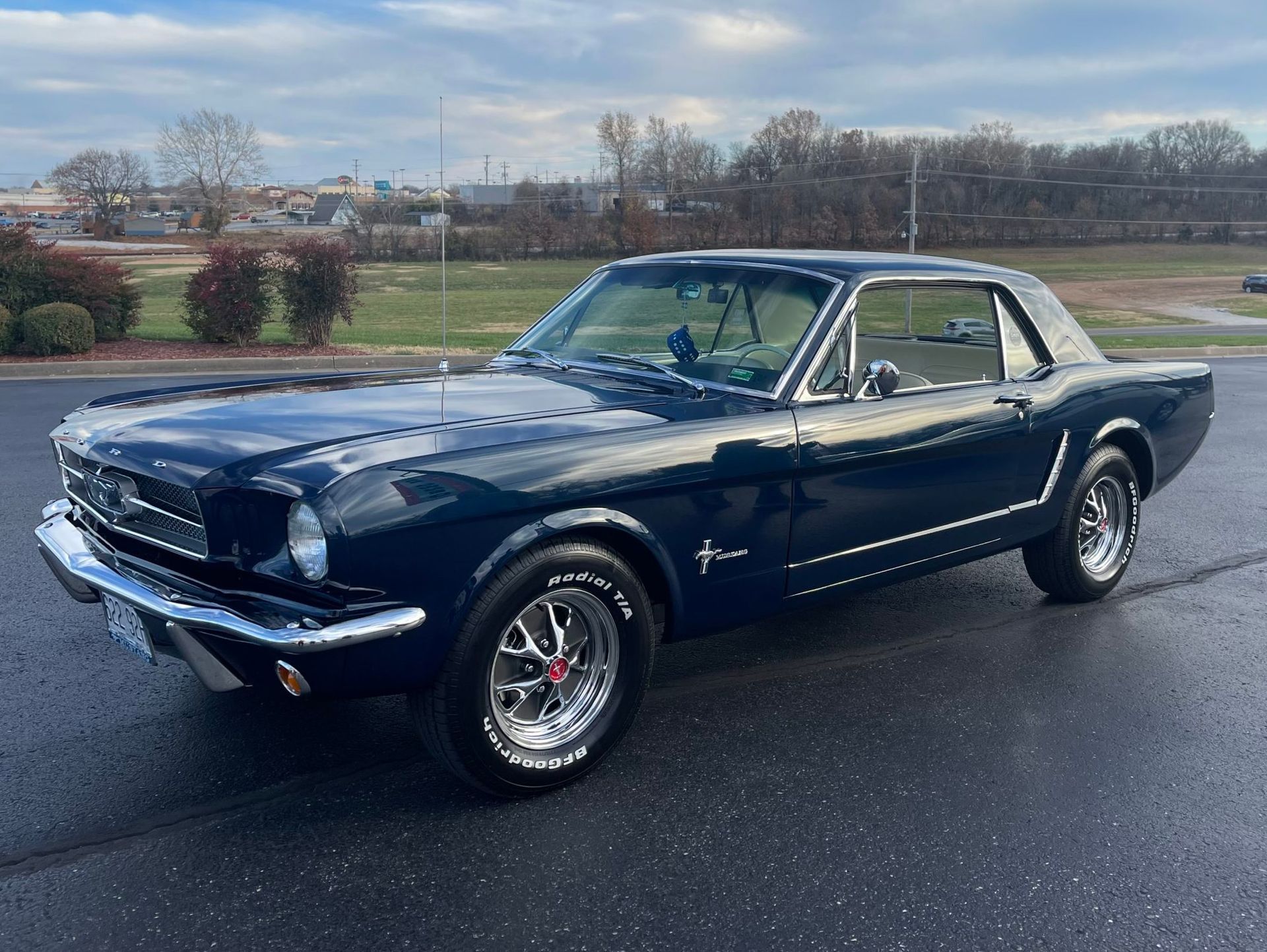 A dark blue classic Ford Mustang coupe parked on an asphalt lot with a cloudy sky and field in the background.