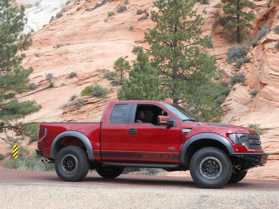 A red Ford Raptor pickup truck parked on a road against a backdrop of rocky, desert cliffs and pine trees.