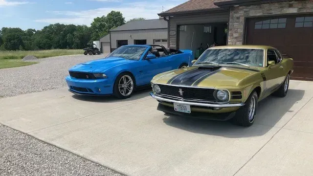 A blue modern Ford Mustang convertible and a vintage gold Mustang parked on a driveway in front of a garage.