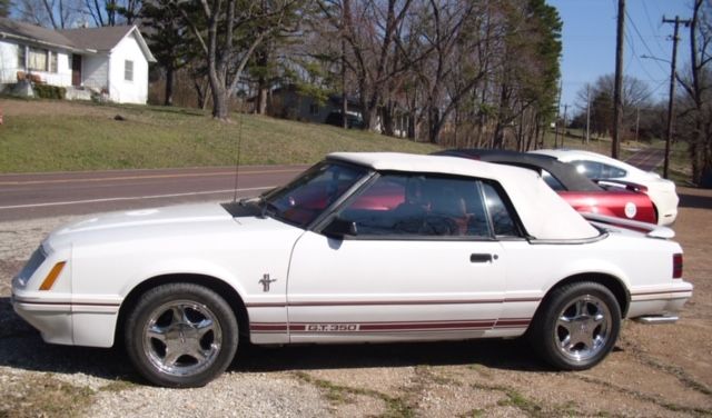 A white Ford Mustang convertible parked on a dirt lot beside a road with a house and trees in the background.