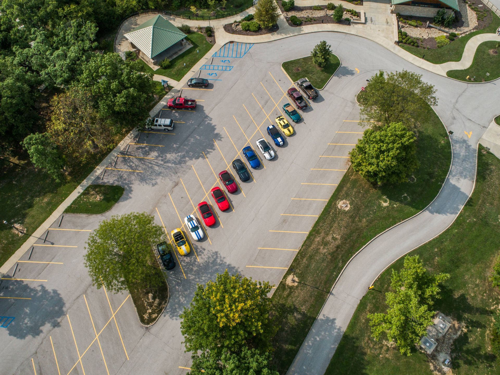 An aerial view of a line of colorful sports cars parked diagonally in a paved lot surrounded by trees and a building.