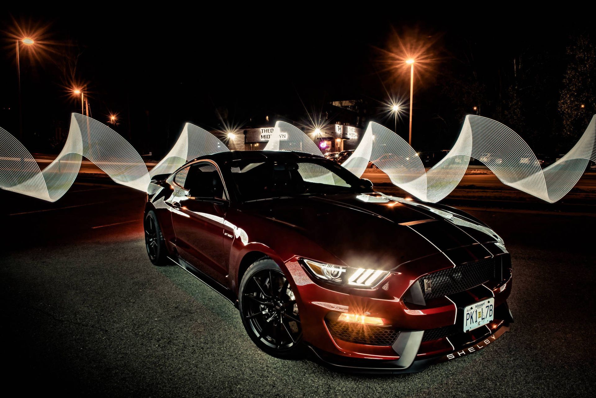 A dark red Ford Mustang parked at night, framed by abstract, swirling light-painted patterns behind it.