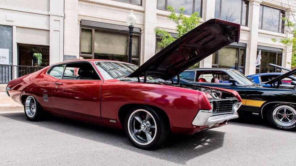 A shiny, deep red vintage muscle car with its hood raised, parked on a city street next to other classic cars.