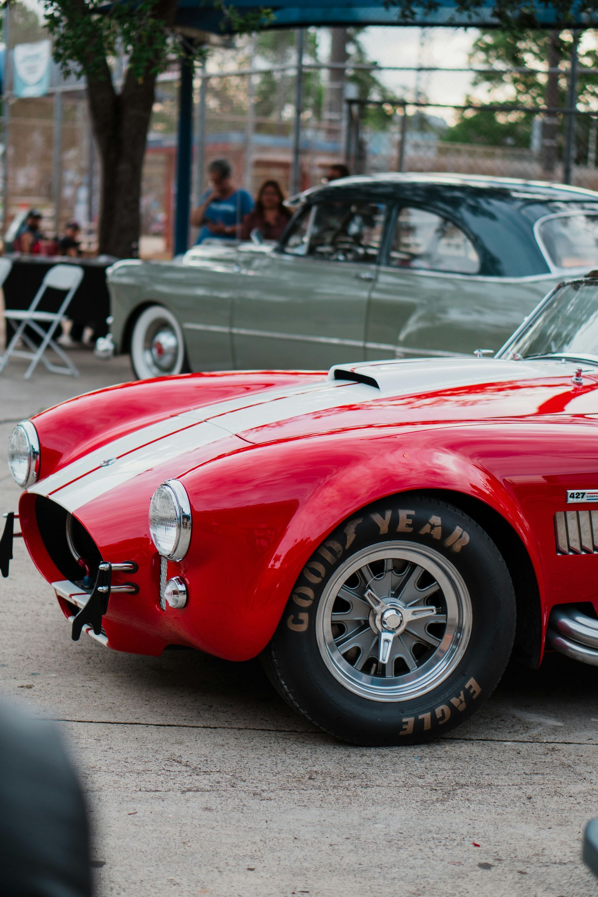 A bright red sports car with a white racing stripe parked next to a classic grey coupe at an outdoor car show.