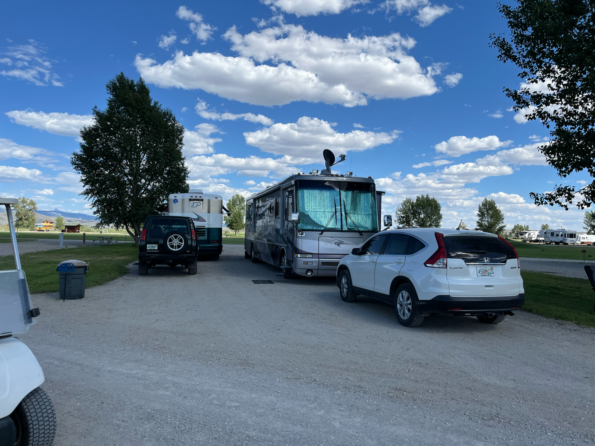 RV parked at a campsite, next to a car and a jeep. Blue sky with clouds overhead.