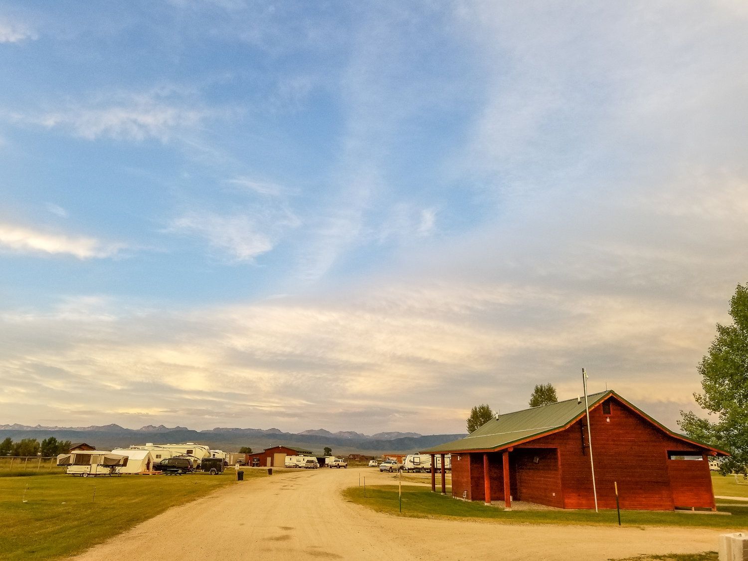Campground with RVs, a brown building, dirt road under a cloudy sky with mountains in the distance.