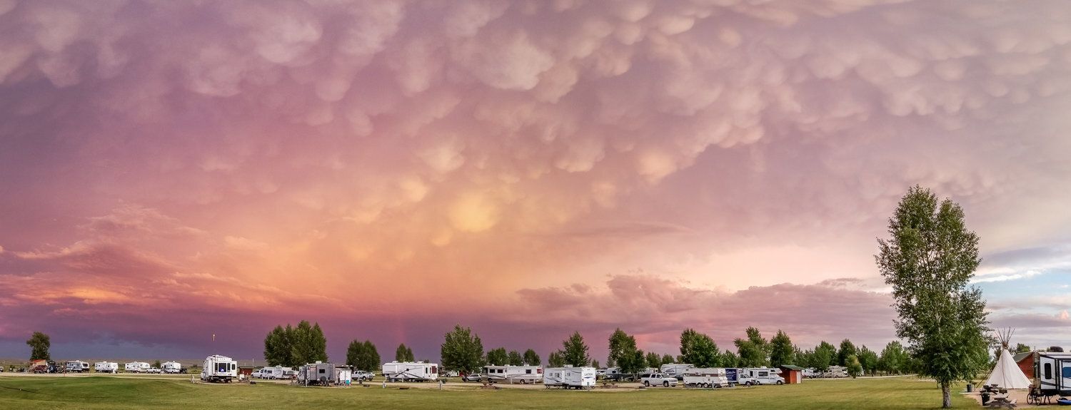 Camping ground under a colorful sunset with clouds. RVs and trees are visible on the green field.