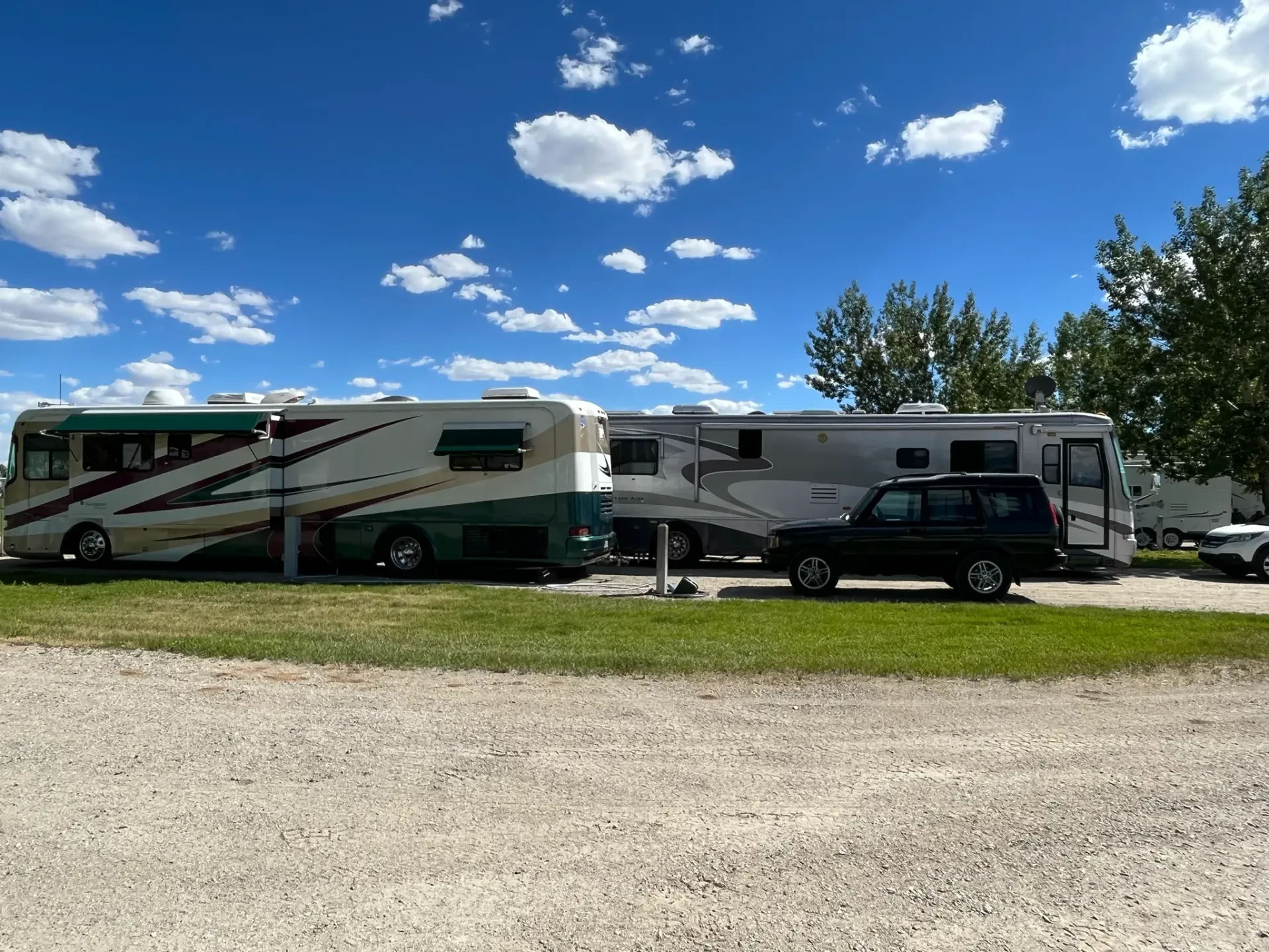RVs parked on gravel under a blue sky with scattered clouds. A black SUV is parked between two of them.