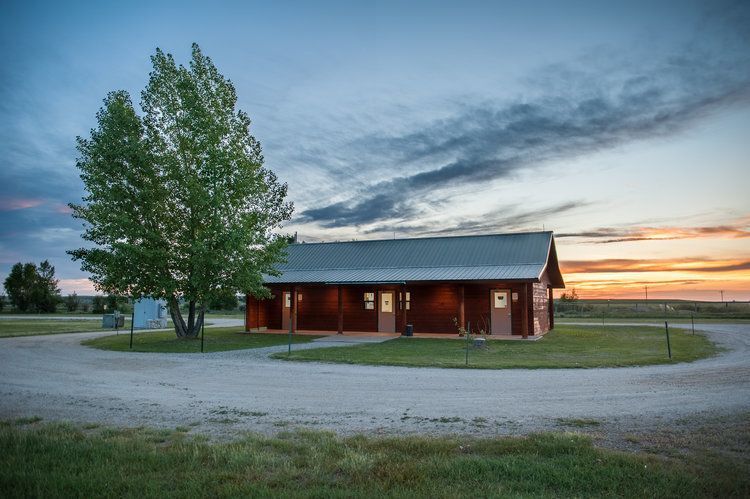 Log cabin under a cloudy sky with a tree in front. Gravel driveway and grassy area around.