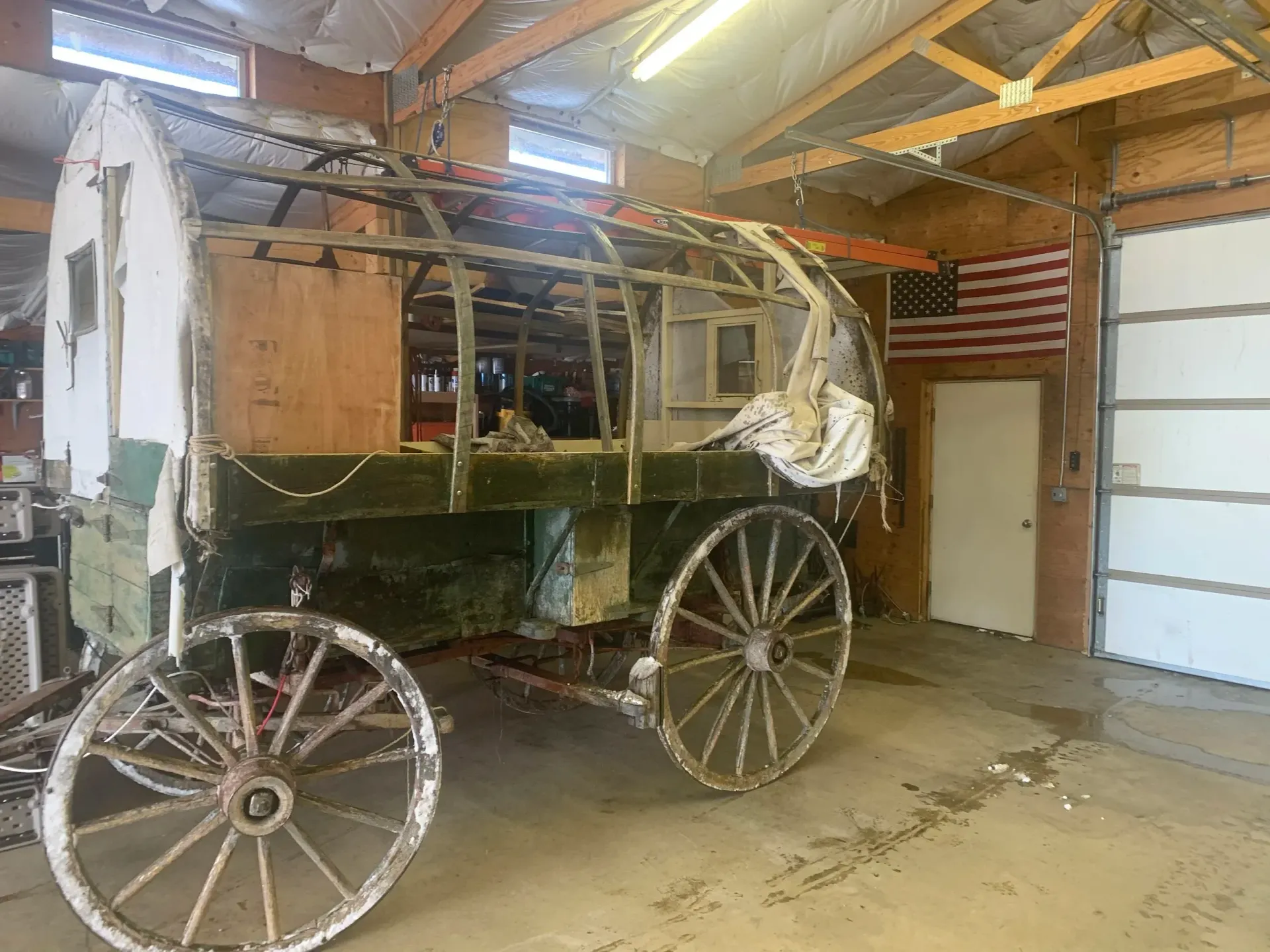A partially restored covered wagon in a garage, with wooden wheels and a green exterior, next to a white door and American flag.