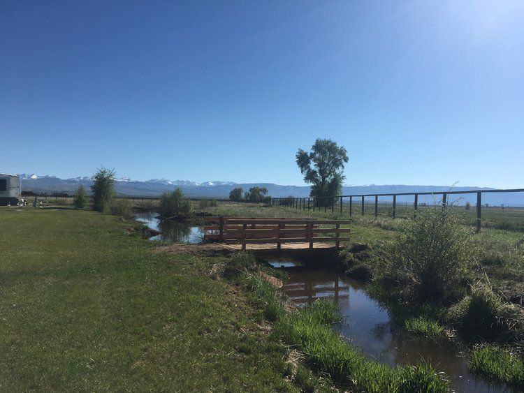 A wooden bridge over a creek in a grassy field, with mountains and a blue sky in the background.
