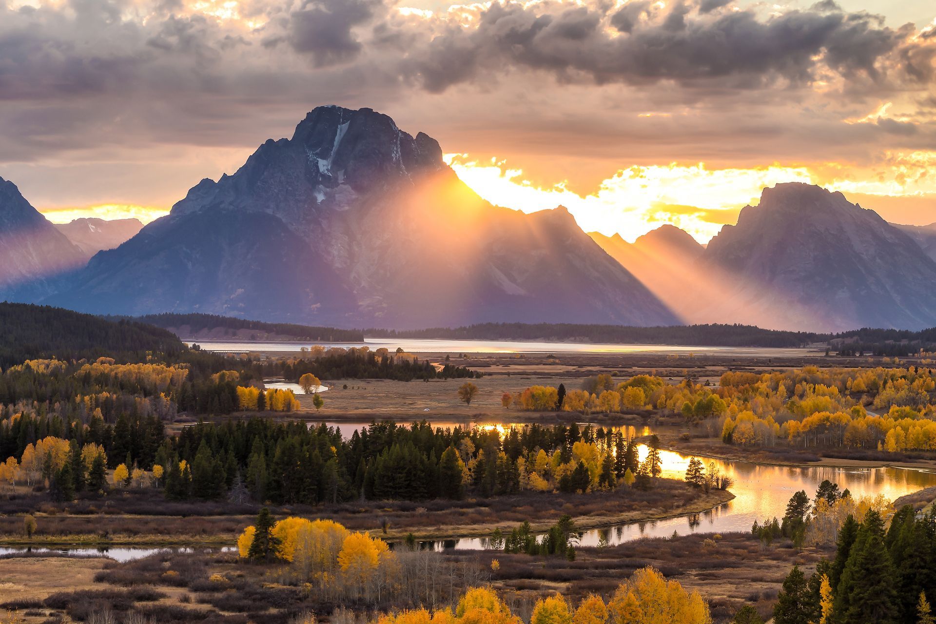 Sun breaking through clouds over a mountain range, reflecting on a lake with autumn foliage.