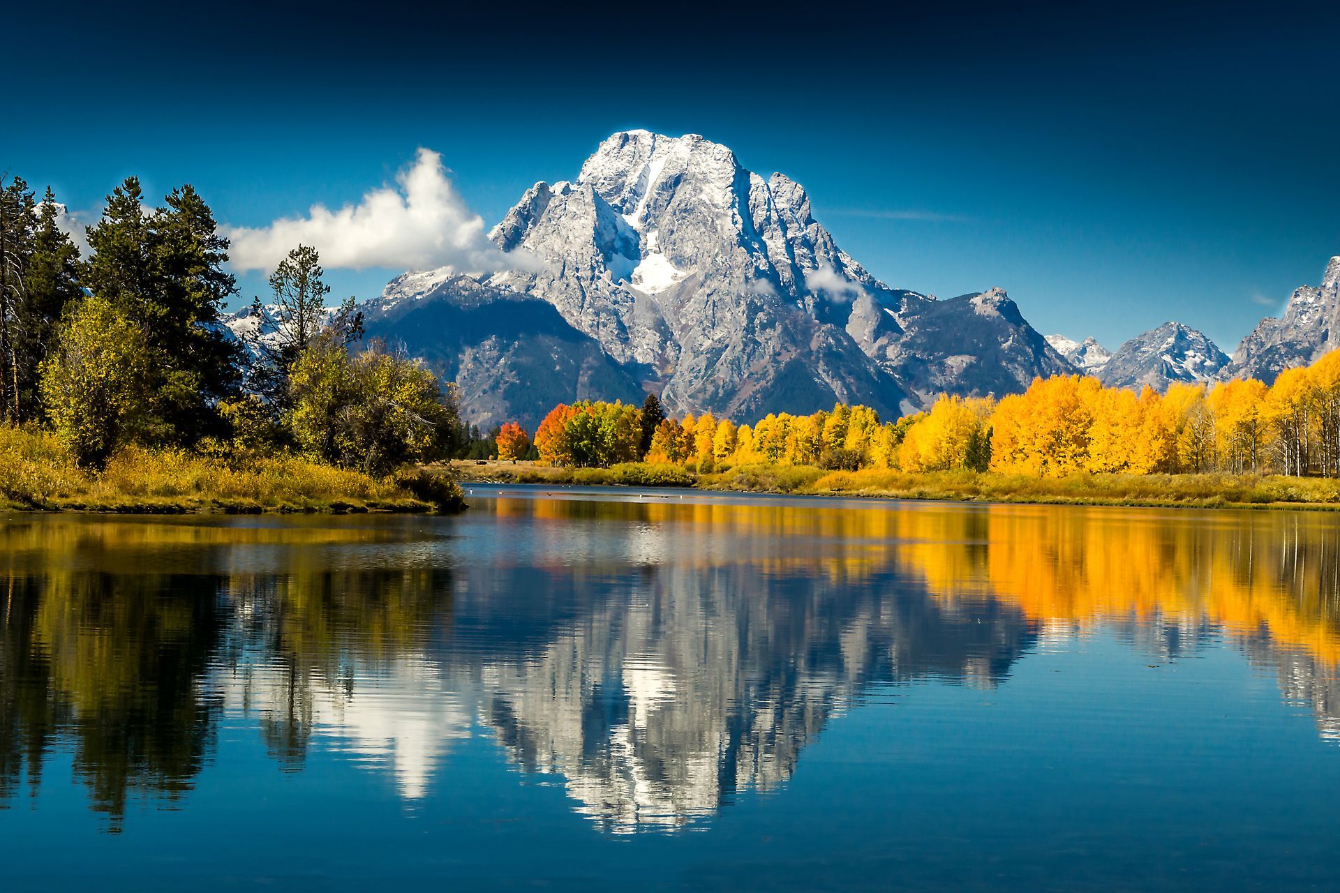 Snow-capped mountain reflected in lake; autumn foliage in foreground; vibrant colors, clear blue sky.