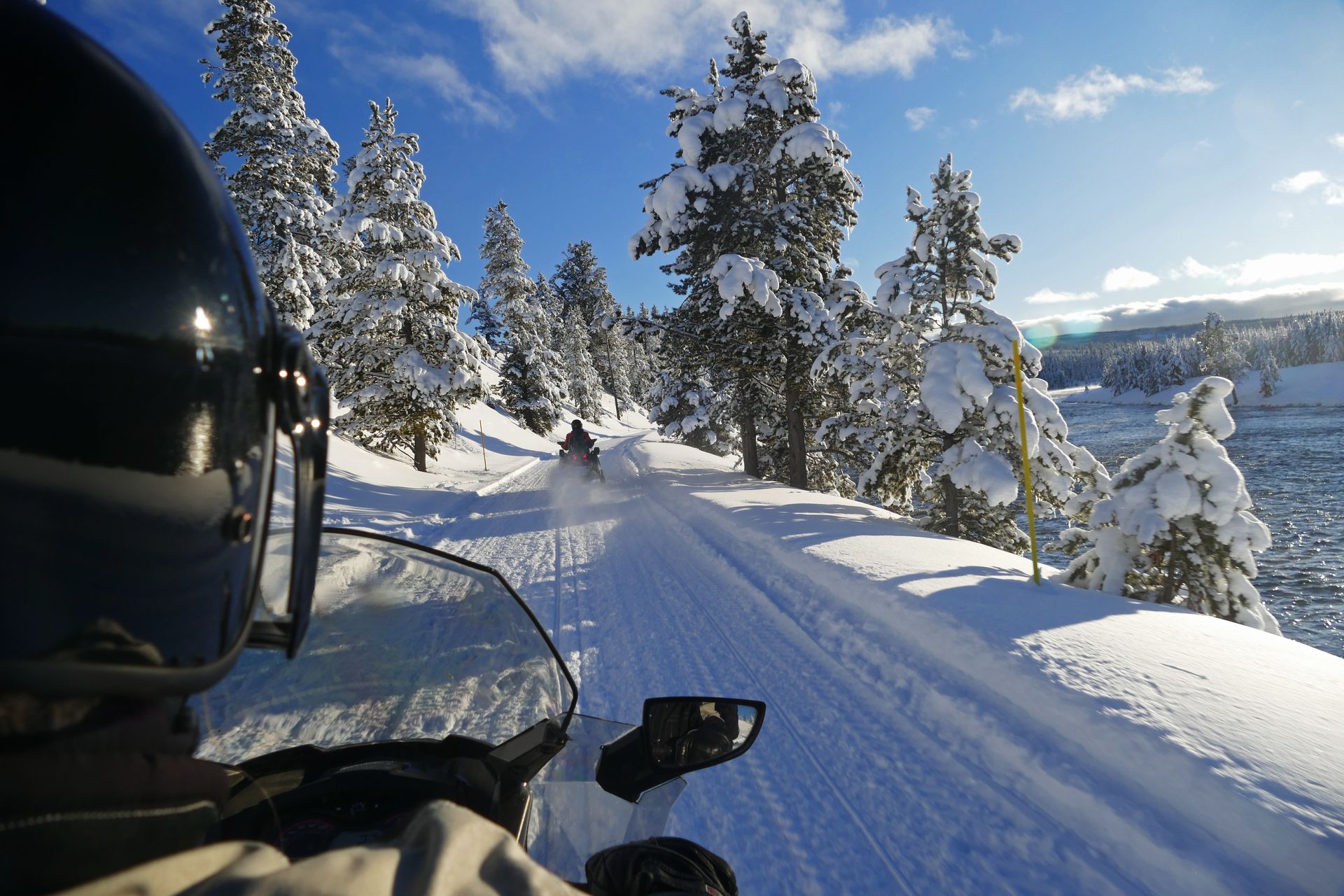 Snowmobiles travel on a snowy trail through a forest on a sunny day.